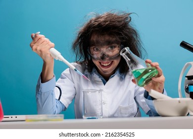 Grinning creepy lunatic silly looking mad scientist with dropper and beaker mixing experimental liquid chemical substances. Crazy chemist with dreadful face expression doing laboratory work.