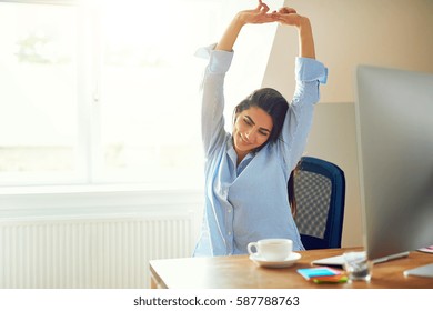 Grinning Young Woman In Blue Long Sleeve Shirt Stretching In Front Of Desk At Home