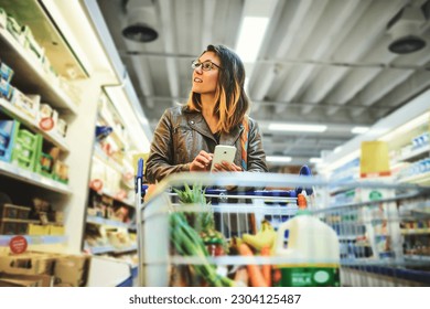 Groceries, phone and woman in supermarket or grocery store using app for shopping choice of food on a shelf. Website, retail and young customer with a decision by a refrigerator with a smartphone