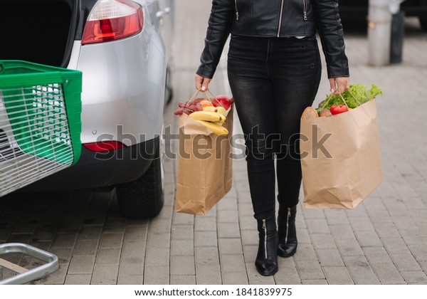 Groceries from a supermarket\
in a eco craft package. Food delivery during quarantine. Paper eco\
bags full of fresh food. Woman hold two package by the car.\
Close-up
