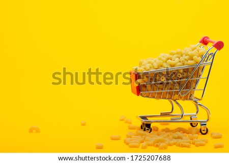 A grocery cart to the top is filled with pasta on a bright orange background, some pasta is lying next to the cart