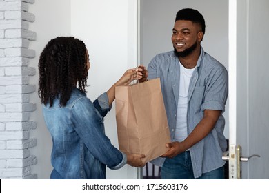 Grocery Delivery. Smiling African American Courier Man Delivering Ordered Goods To Young Woman's Home, Not Wearing Mask