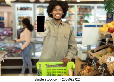 Grocery Shopping App. Black Guy Showing Smartphone With Blank Screen Standing With Shop Cart Full Of Food Products In Supermarket Indoors. Buy Groceries Online. Selective Focus On Phone