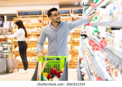 Grocery Shopping. Arabic Man Taking Dairy Product From Shelf In Modern Supermarket. Male Customer Choosing And Buying Food Standing With Shop Cart Full Of Groceries In Store