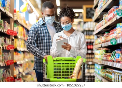 Grocery Shopping. Black Couple Wearing Face Masks In Supermarket, Buying Groceries With Checklist, Walking With Trolley In Food Store Indoors. Customers Buy Essentials During Virus Pandemic Outbreak