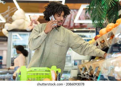 Grocery Shopping. Black Guy Talking On Phone While Choosing Fruits Standing With Shop Cart Buying Food Products In Modern Supermarket Indoors. Consumerism Lifestyle Concept