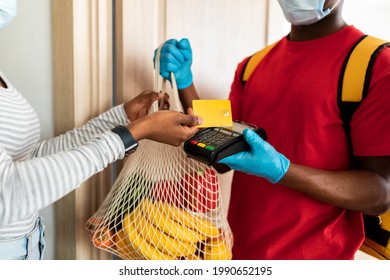 Grocery Shopping Delivery. Black Female Buyer Paying Courier With Credit Card For Groceries Products From Supermarket Standing In Doorway At Home. Man In Mask Delivering Food To Customer. Cropped