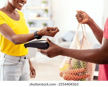 Grocery Shopping Delivery. Black Lady Paying With Smartwatch For Groceries Receiving Shopper Bag With Food Products From Supermarket Courier Guy At Home. Cropped Shot, Selective Focus - Powered by Shutterstock