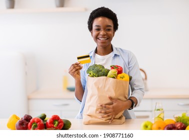 Grocery Shopping. Happy African Woman Holding Credit Card And Bag Full Of Vegetables Standing Posing In Kitchen At Home, Smiling To Camera. Household, Healthy Food And Nutrition Concept