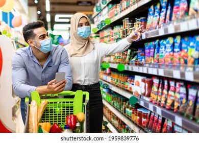 Grocery Shopping. Muslim Couple Choosing Food In Supermarket, Arabic Wife In Hijab And Protective Face Mask Pointing Finger On Shelf With Products. Islamic Family Purchasing Essentials In Store