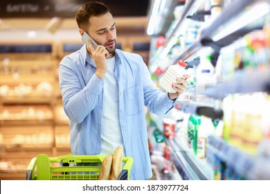 Grocery Shopping. Portrait Of Male Customer Holding Bottle Of Milk Or Yoghurt, Talking On Smartphone, Asking Wife About List Of Purchases, Standing Near Fridge With Dairy Products