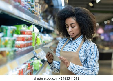 A grocery store employee in an apron checks the inventory on the shelves using a tablet. The worker appears focused and attentive, ensuring the store is well-stocked and organized for customers. - Powered by Shutterstock