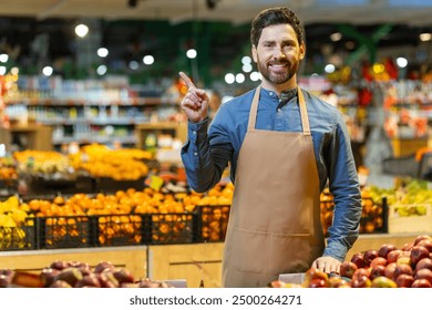 Grocery store employee in apron standing in produce section pointing finger. Bright fruits and vegetables on display creating colorful background. Image of customer service, retail, food, and market. - Powered by Shutterstock