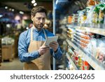 Grocery store employee with apron using tablet for inventory management in food aisle. Worker checking stock and organizing products in supermarket. Efficient retail operations with technology