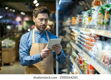 Grocery store employee with apron using tablet for inventory management in food aisle. Worker checking stock and organizing products in supermarket. Efficient retail operations with technology - Powered by Shutterstock