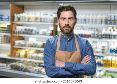 Grocery store employee stands confidently in front of dairy aisle. Wearing blue shirt and apron, he demonstrates professionalism in retail setting. Background includes various products on shelves. - Powered by Shutterstock