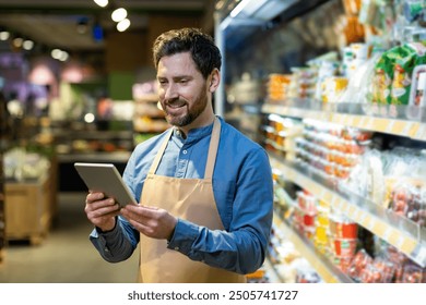 Grocery store employee wearing apron stands in supermarket aisle using tablet. Man checks inventory and manages tasks on device. Shelves filled with packaged goods in background - Powered by Shutterstock