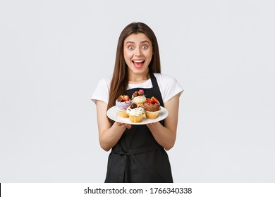Grocery store employees, small business and coffee shops concept. Excited female barista, cafe working in black apron holding plate with cupcakes, showing new menu summer desserts in cafe