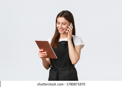 Grocery Store Employees, Small Business And Coffee Shops Concept. Cute Smiling Saleswoman In Black Apron Confirm Order, Talking To Customer On Phone And Looking At Digital Tablet