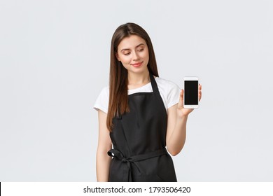 Grocery store employees, small business and coffee shops concept. Pretty young female cafe worker in black apron advertise mobile application, showing smartphone screen, stand white background - Powered by Shutterstock