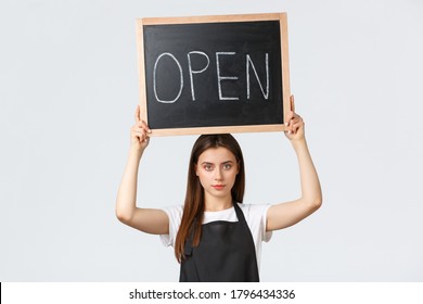 Grocery Store Employees, Small Business And Coffee Shops Concept. Serious-looking Young Barista In Black Apron Raise Open Sign, Inform Customers That Shop Or Cafe Working After Covid Pandemic