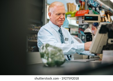 Grocery Store Manager Sitting At Checkout Counter And Looking At Something On Computer. Senior Man Working At Supermarket Checkout Counter.