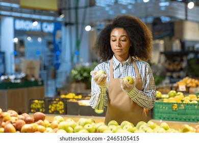 Grocery store, a woman wearing an apron, gloves carefully inspects and organizes apples. Shelves and produce can be seen in the background, showcasing a professional worker in a busy supermarket - Powered by Shutterstock