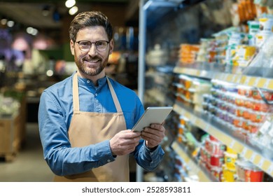 Grocery store worker in apron smiling while using digital tablet for inventory management. Enhancing customer service with technology in retail setting. Efficient and friendly service - Powered by Shutterstock