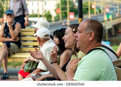 Grodno, Belarus July 10, 2022: An Elderly Man With His Family Is Resting In The Park Sitting On A Bench. A Pensioner With His Granddaughter At An Outdoor Concert. The Man Smiles Cheerfully