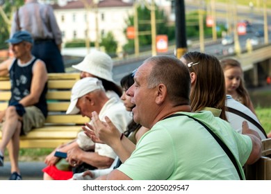 Grodno, Belarus July 10, 2022: An Elderly Man With His Family Is Resting In The Park Sitting On A Bench. A Pensioner With His Granddaughter At An Outdoor Concert. The Man Smiles Cheerfully