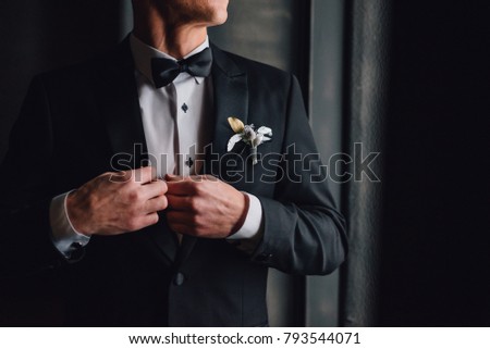 Groom in black tuxedo and bowtie correct his buttons on white shirt. Wedding. Details