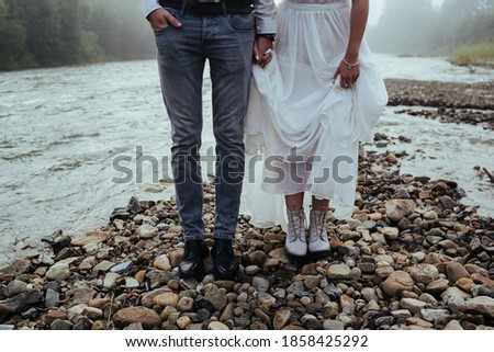 The groom and the bride in a white long dress stand holding hands in front of a river on an stone coast. wedding shoes