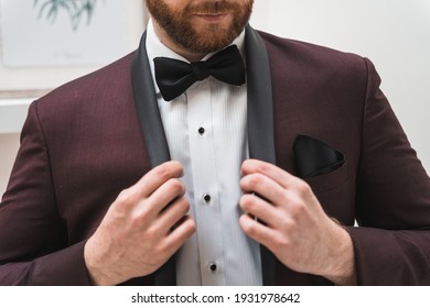A groom buttons his maroon tuxedo jacket as he prepares for his wedding day