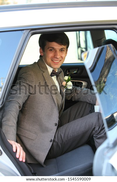 Groom in a car in the back seat in a gray retro\
suit and smiling