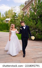 The Groom In A Classic Suit And The Blonde Bride In A White Dress Walk Together Holding Hands. The Concept Of Love, Mutual Understanding And Happy Family Life.