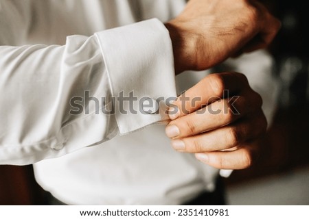 The groom fastens the cufflinks at the wedding reception with a large plon. The man wears a white shirt. The groom is preparing for the wedding