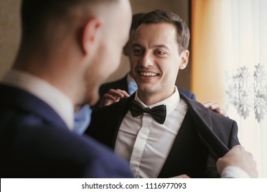 Groom Getting Ready In The Morning With Groomsmen In The Room. Man Hands Putting On Jacket And Bow Tie, Standing At Window Light In Hotel. Preparation For Wedding Day