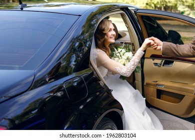 Groom Helps Happy Young Bride To Get Out Of Wedding Car. Elegant Bride Holds Wedding Bouquet Of White Roses In Her Hands. The Bride And Groom Arrived At The Wedding Ceremony