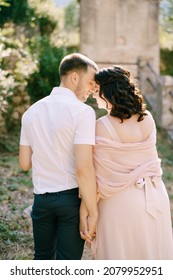 Groom Hugs Bride Against The Backdrop Of The Building In The Garden. Back View