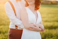 The Groom Hugs Bride From Behind. Woman And Man In Wedding Dress Outdoors Holding Hands. A Young Man Gently Embraces His Bride In A Wedding Dress. Wife And Husband In The Field. Chaplet