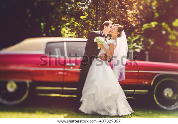 Groom hugs bride\'s waist kissing her in a cheek\
before old red Dodge