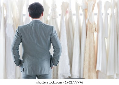 The groom man in a gray suit stands and looks back at the bride's dress. A man standing to help bride fitting choosing wedding dress in studio. Concept happy choose for bride in best day
