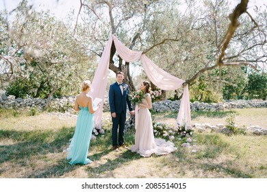 Groom stands with bride near a tree in a green grove in front of the wedding ceremony master - Powered by Shutterstock