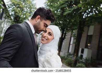 groom in suit hugging cheerful muslim bride in hijab and white dress
