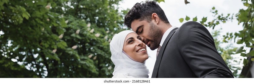 groom in suit hugging cheerful muslim bride in white hijab, banner