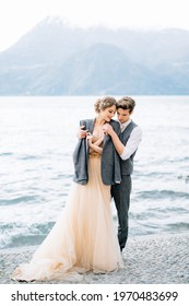 Groom threw a jacket over the shoulders of bride hugging her on the embankment of Lake Como near the water
