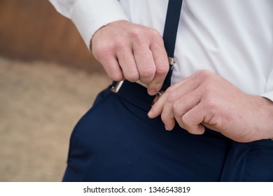 Groom wearing white shirt and navy pants fastening suspenders to pants