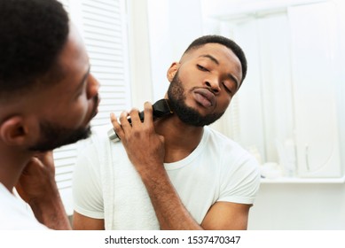 Grooming Concept. Young Black Man Looking At Mirror And Shaving Beard With Trimmer Or Electric Shaver At Bathroom, Free Space