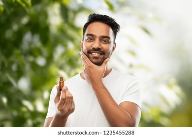 grooming and people concept - smiling young indian man applying lotion or beard oil over green natural background