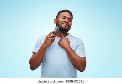 grooming and people concept - smiling young african american man shaving beard with trimmer over blue background